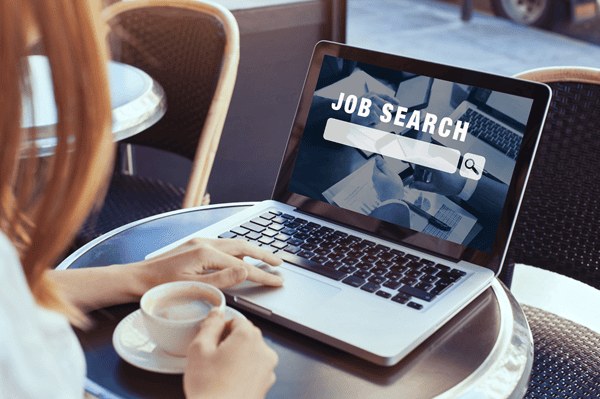 Eine Person von der Seite mit einer Tasse in der Hand, vor Ihr auf einem runden Tisch ein Laptop, auf dessen Bildschirm steht "JOB SEARCH"