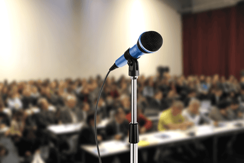 Im Vordergrund ein Standmikrofon, im Hintergrund eine Menschmenge sitzender Personen