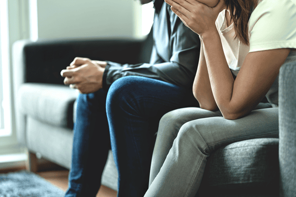 Zwei Personen Sitzen auf einem Sofa, seitliche Perspektive, die Person die links sitzt hält beide Hände an den Mund, die Köpfe sind nicht zu sehen