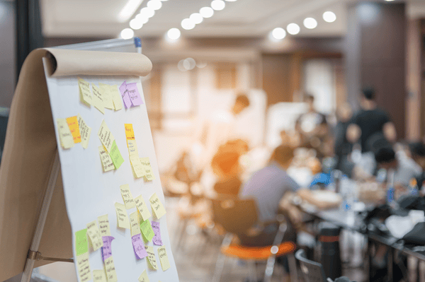 Ein Flipchart vorne links im Bild mit beschrifteten Haftzetteln, im Hintergrund Menschen die um einen Tisch herum stehen und sitzen