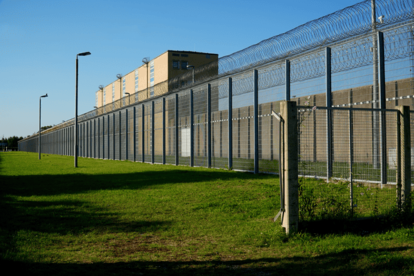 Ein hoher Zaun, obenauf mit Stacheldraht, dahinter eine großes Gebäude