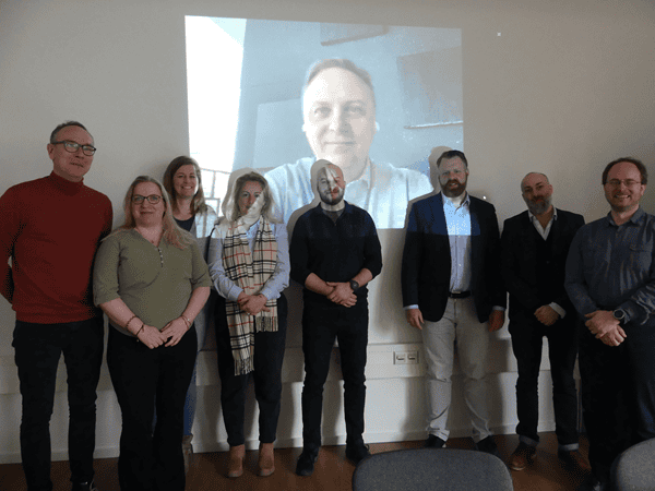 Acht Personen, in Gänze abgebildet, stehen nebeneinander und schauen in die Kamera, im Hintergrund die Projektion einer weiteren Person