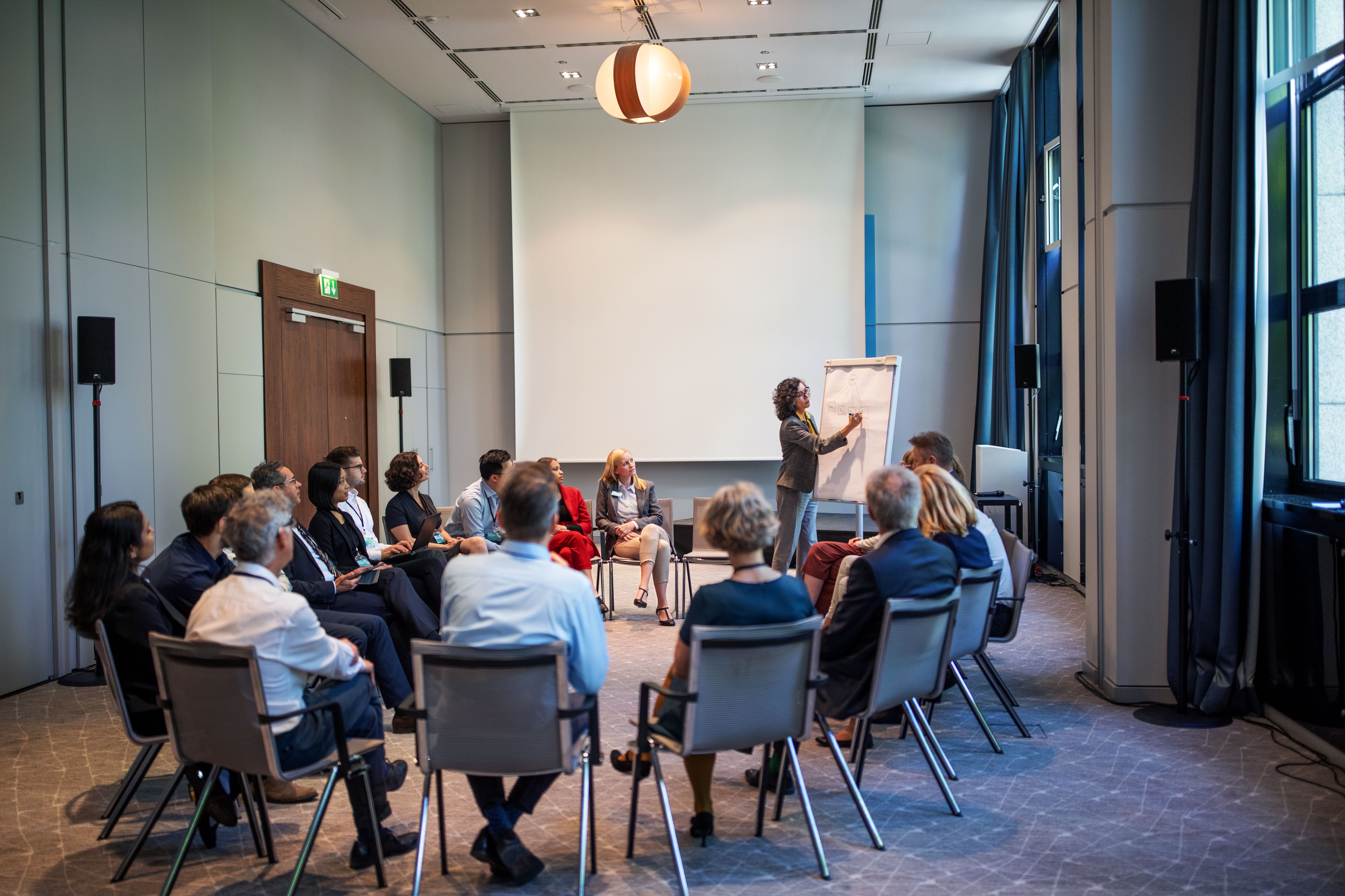 Mehrere Personen sitzen in einem Zimmer in einem Stuhlkreis und blicken in Richtung einer stehenden Person, die in Richtung eines Flipcharts gerichtet ist