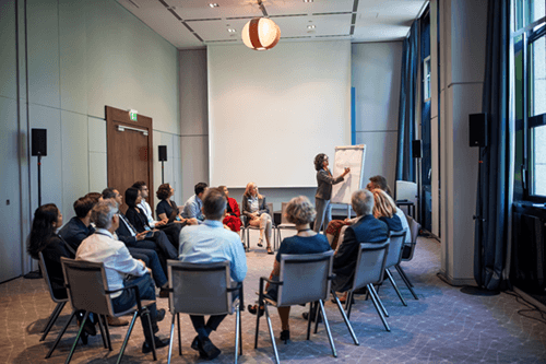 Mehrere Personen sitzen in einem Zimmer in einem Stuhlkreis und blicken in Richtung einer stehenden Person, die in Richtung eines Flipcharts gerichtet ist