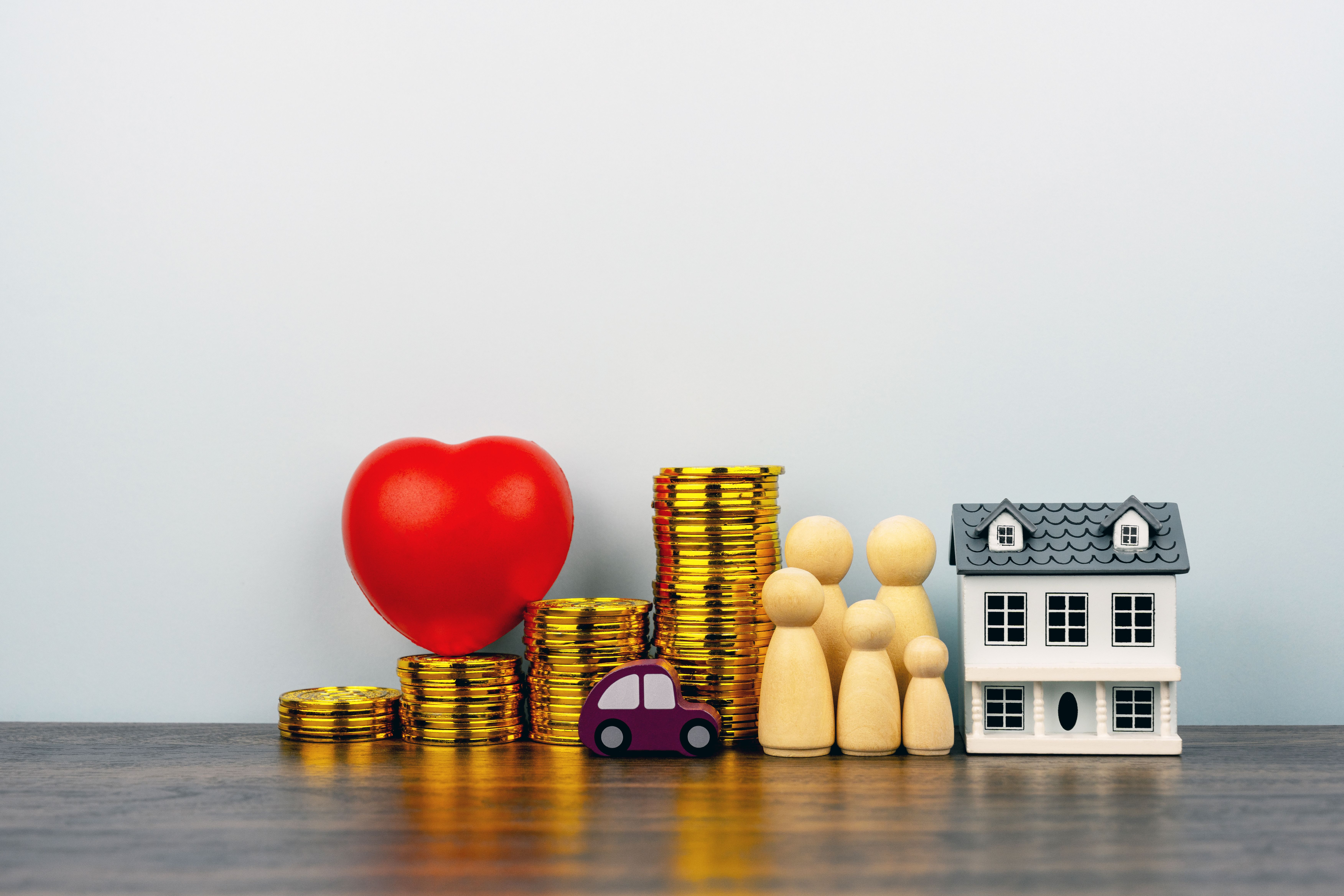 Ein Miniaturhaus, daneben Miniaturfiguren, ein Miniaturauto, Stapel von Goldmünzen und ein rotes Herz