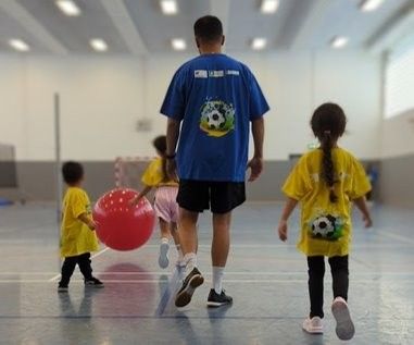 Kinder mit gelben T-Shirts, ein Mann mit einem blauen T-Shirts, in einer Sporthallte mit einem roten Ball