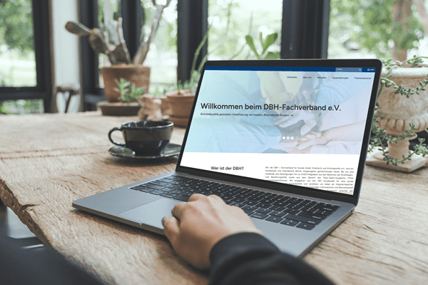 Ein aufgeklappter Laptop mit der Startseite des DBH-Webseite auf dem Bildschirm, er steht auf einem Holztisch, im Hintergrund sind Pflanzen zu sehen